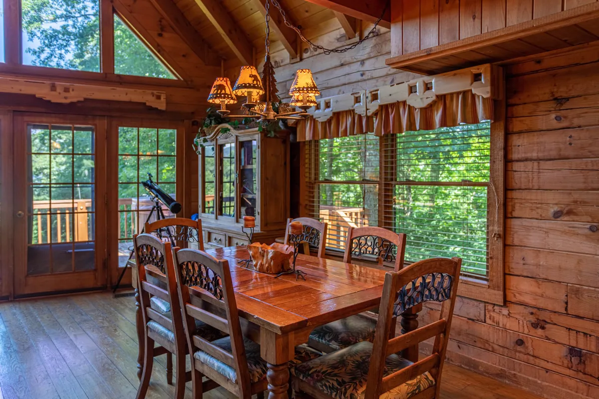 A welcoming dining area featuring a wooden dining table set beneath a chandelier, with expansive windows revealing nature views beyond. Wooden cabinets add rustic charm, and the space feels ideal for both intimate dinners and lively gatherings.