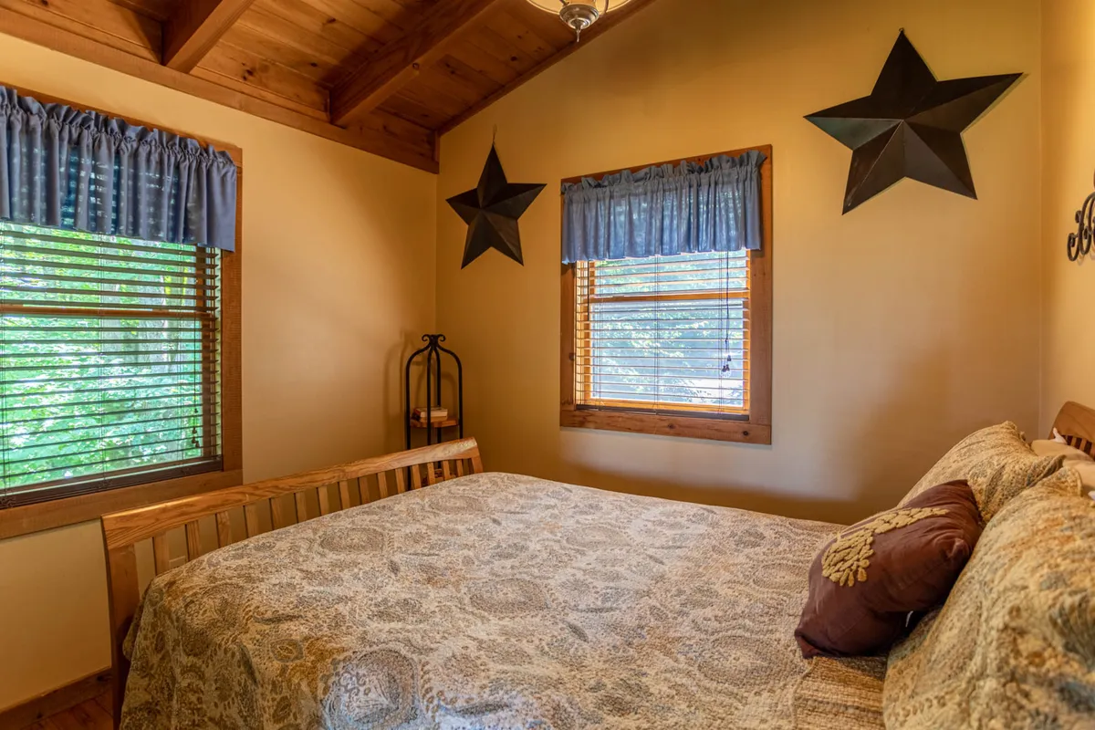 A warmly lit bedroom with a bed covered in patterned bedding, wooden ceiling beams, decorative star wall hangings, and windows dressed with blue curtains. The room is inviting and filled with natural light, emphasizing comfort and solitude.