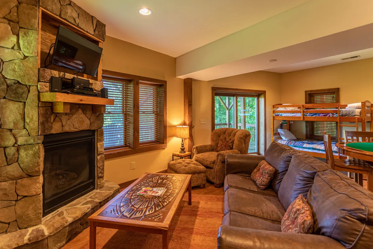 A cozy lower-level recreation room featuring a stone fireplace as the focal point, flanked by comfortable seating, bunk beds for kids, and a wooden coffee table. The room radiates warmth and is designed for relaxation, family gatherings, game nights, and watching movies. The inviting ambiance is enhanced by wood accents and soft lighting.