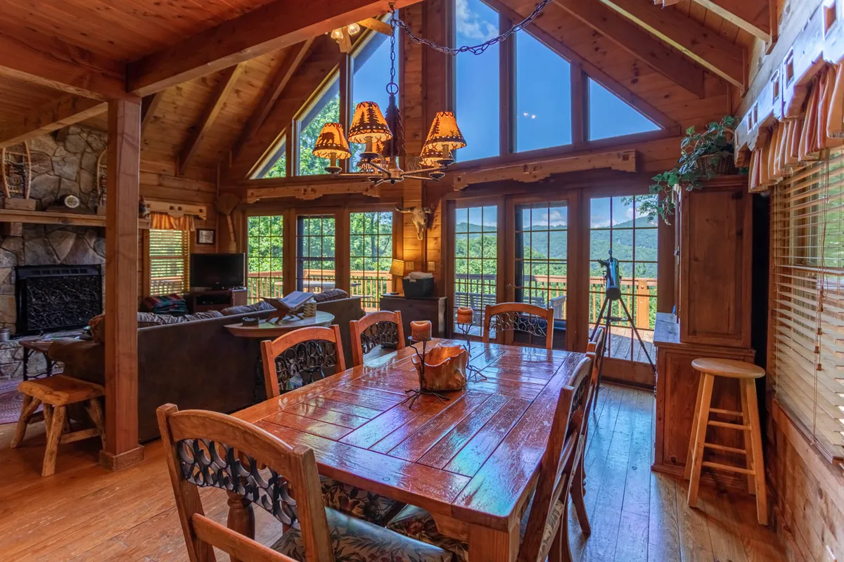 This image shows a spacious dining area centered around a large wooden table, with a striking stone fireplace and oversized windows that offer views of the surrounding mountains. The warm lighting and inviting decor evoke a sense of togetherness, making it ideal for family meals, game nights, or simply gathering to enjoy the ambiance and vistas.