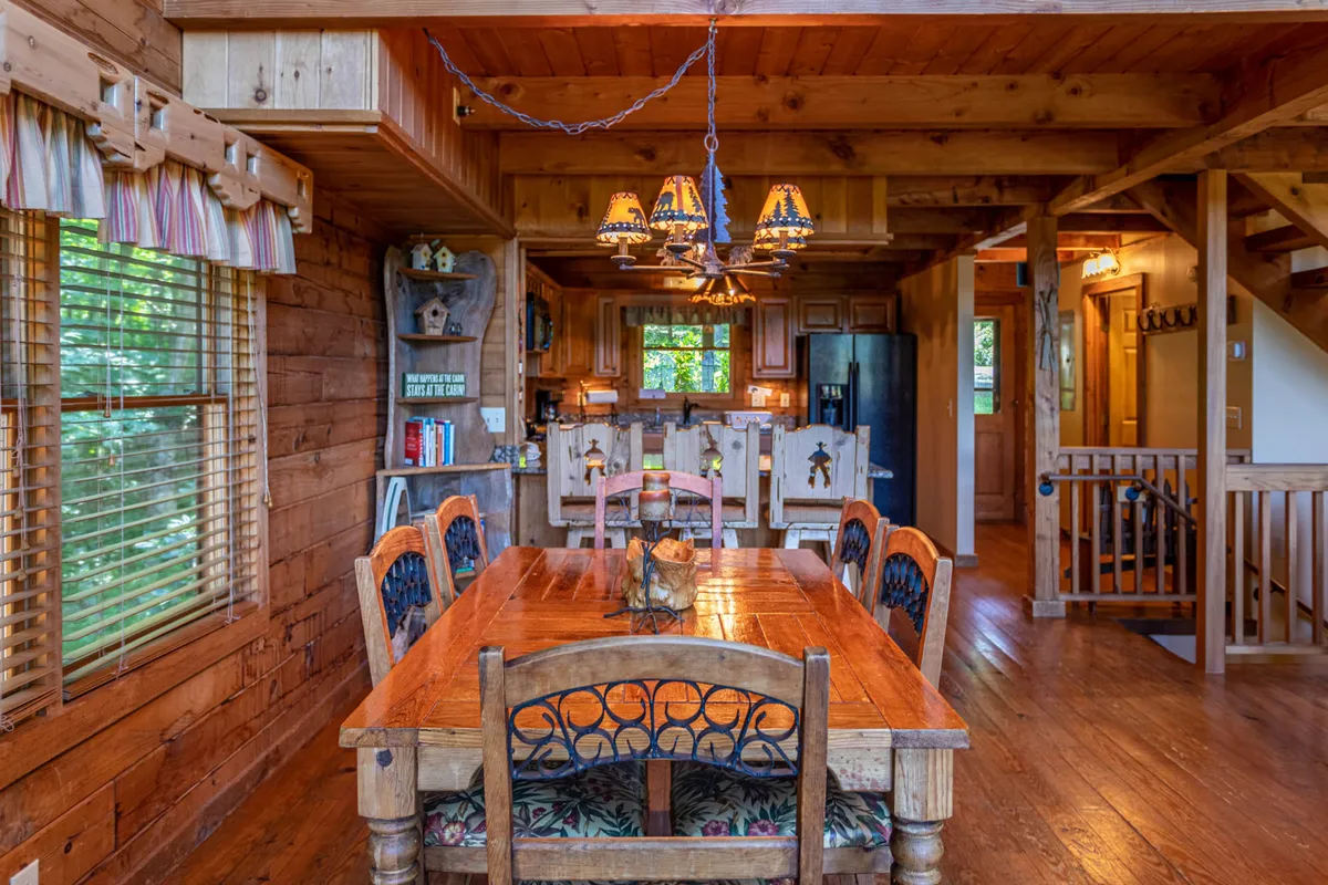 A spacious, welcoming dining area featuring a large wooden table under a rustic chandelier, with exposed beams and an adjoining kitchen. The scene suggests a flexible space ideal for group gatherings, game nights, or collaborative work sessions, with ample natural light and warmth.