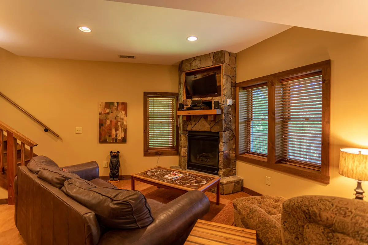 A cozy living area featuring a stone fireplace, inviting sofa, rustic coffee table, and large windows that let in natural light. The windows offer glimpses of the outdoors, suggesting a connection to the cabin's scenic mountain environment. The setup is ideal for relaxing after a day of exploring seasonal activities, with options for enjoying a warm fire, reading, or gathering for board games.