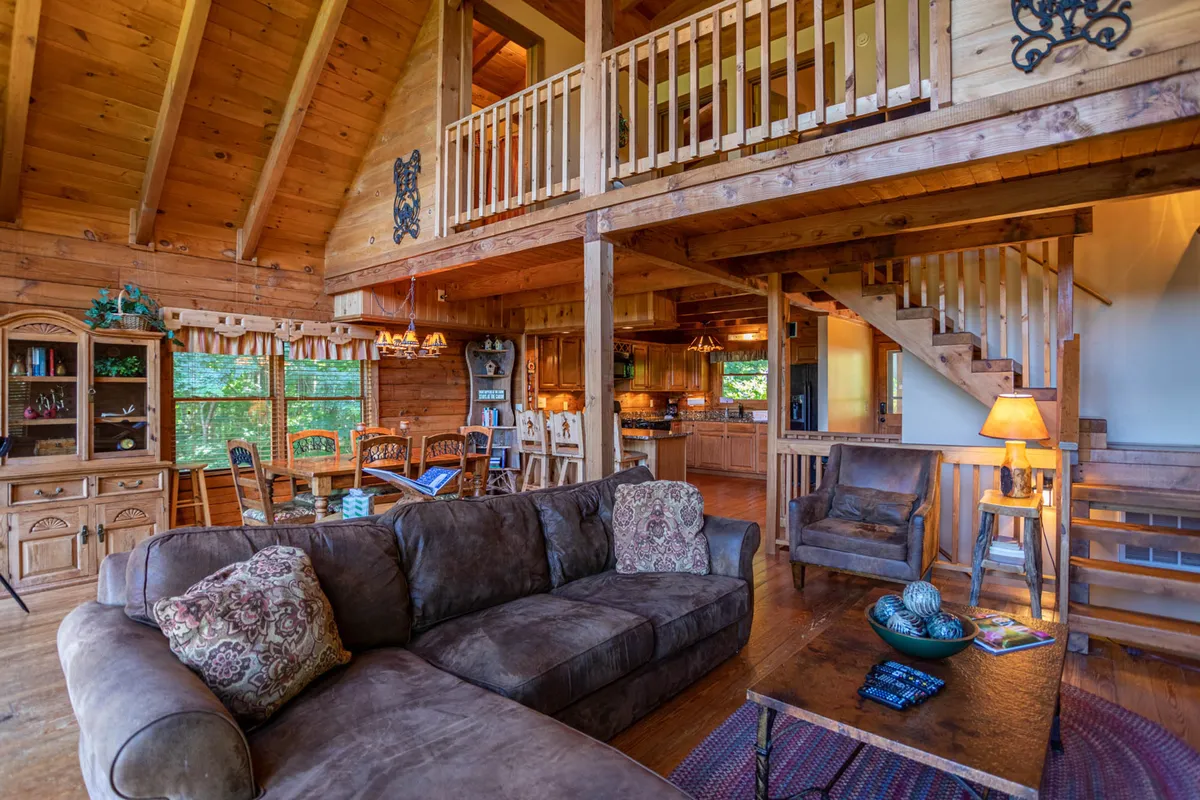 A comfortable, homely open-concept space with a large brown sectional sofa, a wooden dining table, staircase to a loft, and visible kitchen cabinetry. The area feels spacious yet cozy, perfect for gatherings and everyday living.