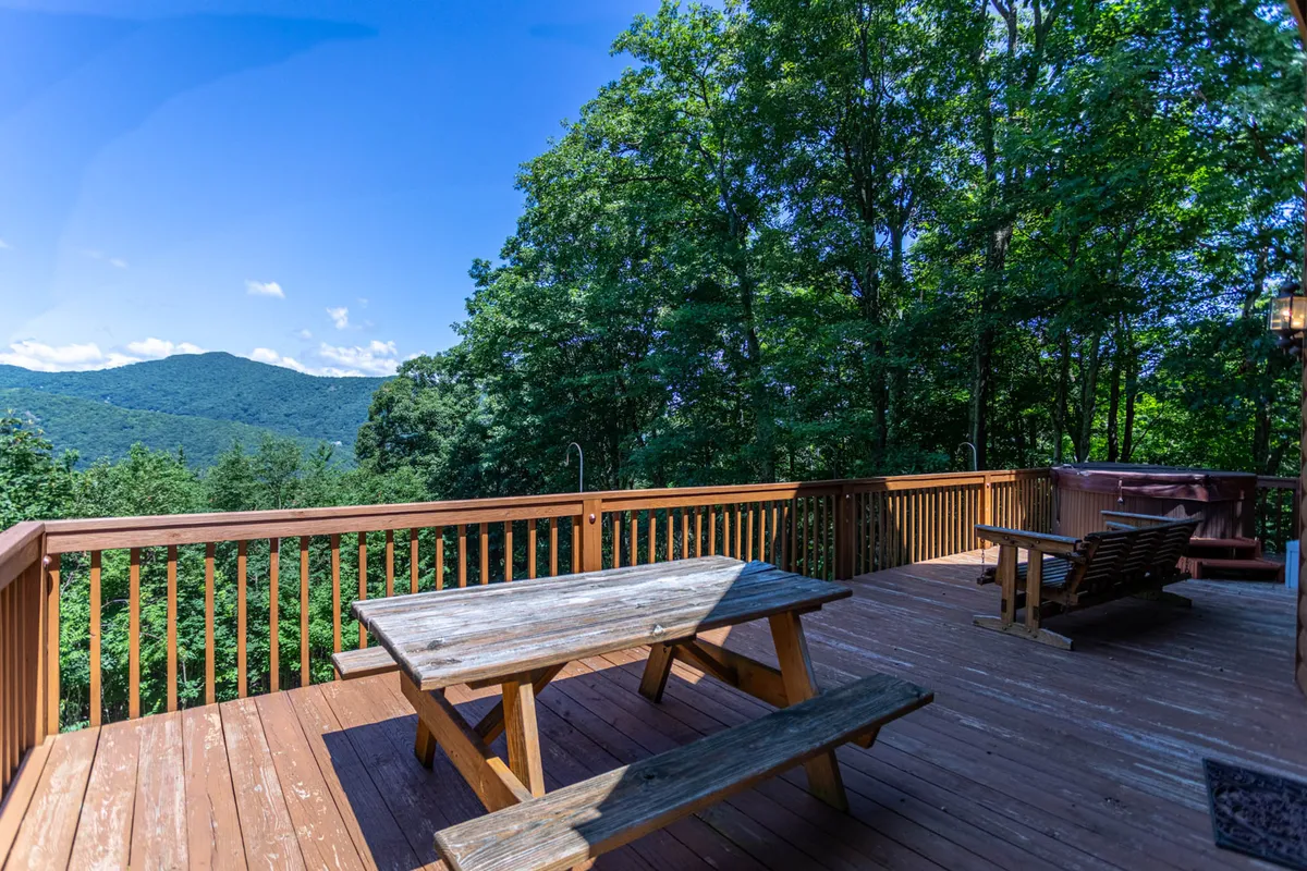 An exterior shot of the lower-level deck with wooden flooring, a picnic table, and a hot tub, all overlooking a stunning mountain view. The deck feels inviting and is perfect for outdoor dining, relaxing, or gathering while enjoying nature's beauty.