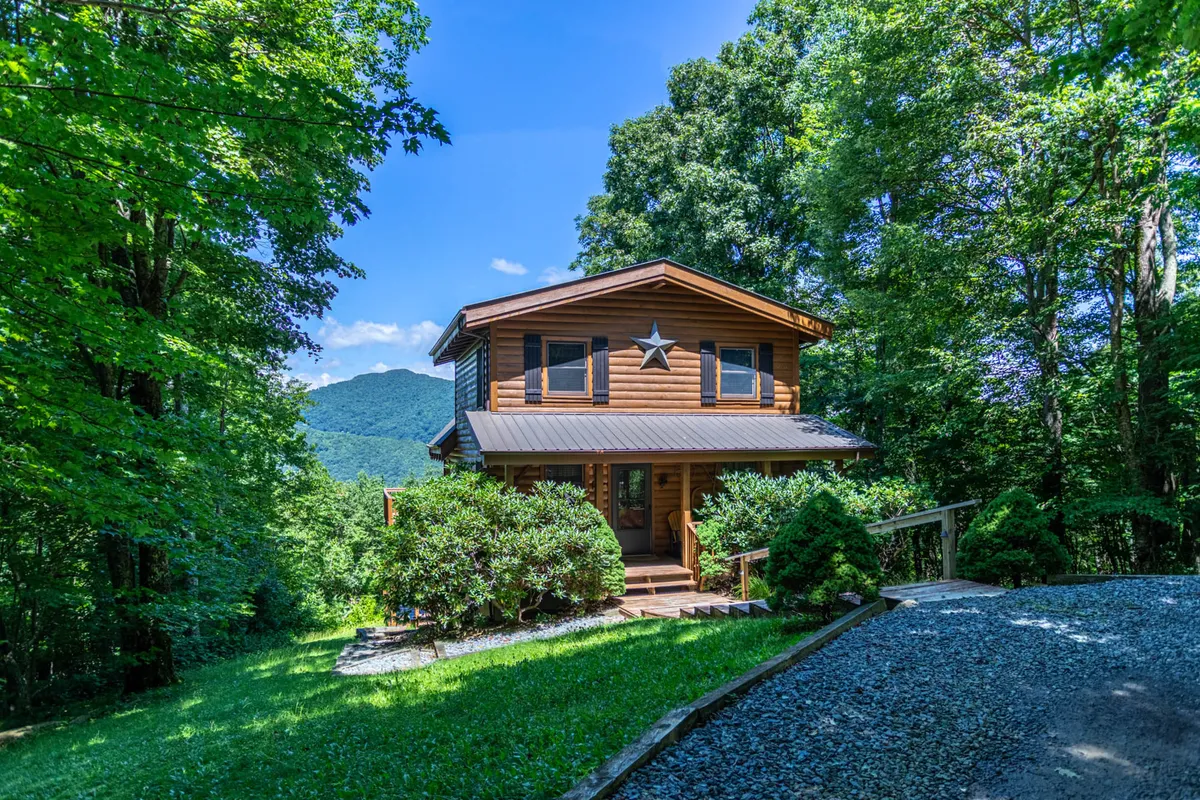 A charming wooden cabin nestled amidst lush landscaping, with expansive mountain views unfolding beyond the porch. The scene is bathed in the soft glow of sunset, inviting guests to relax outdoors and soak in the surrounding natural beauty.