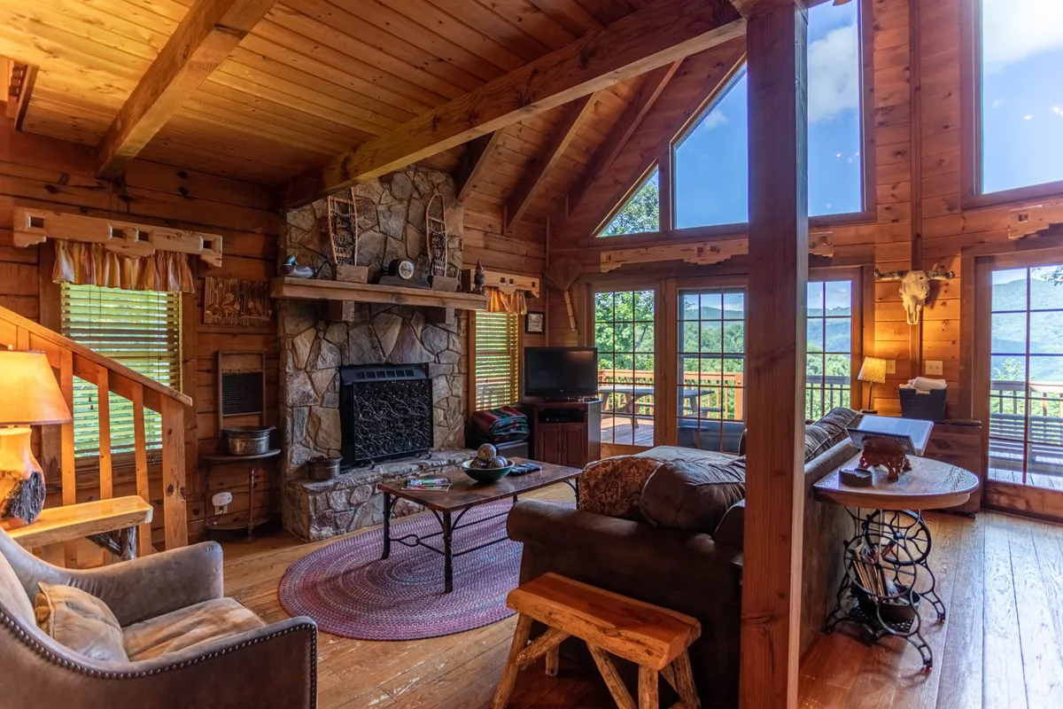 A warm and welcoming living area featuring a stone fireplace, large windows letting in mountain views, wooden beams, and comfortable seating. The atmosphere invites relaxation and gathering, with ample space for pets and people alike.