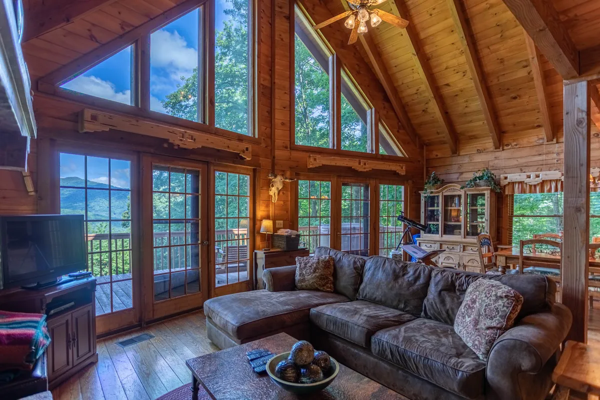 This image features a cozy living room with a large sectional sofa facing expansive windows that offer panoramic mountain views. The space showcases rustic wooden ceiling beams and inviting decor, immersing guests in warmth and natural light. The living area is arranged for family gatherings and relaxation, with the outdoors always in view.