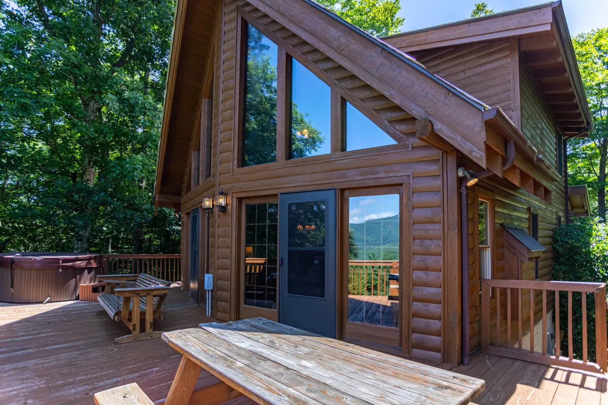 This exterior image features a spacious wooden deck with a hot tub, large windows looking into the cabin, and a dining table set for outdoor meals. The deck is surrounded by lush greenery, creating a seamless transition between indoor comforts and the natural mountain environment.