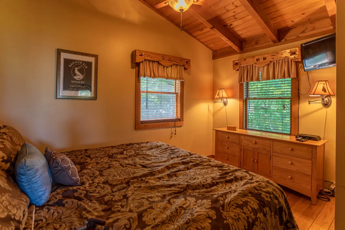 A spacious bedroom featuring a bed with ornate bedding, a wooden dresser, a window with blinds, and a wall-mounted TV. Decorative framing on the windows adds character. The scene is calm and comfortable, with natural light filtering in.
